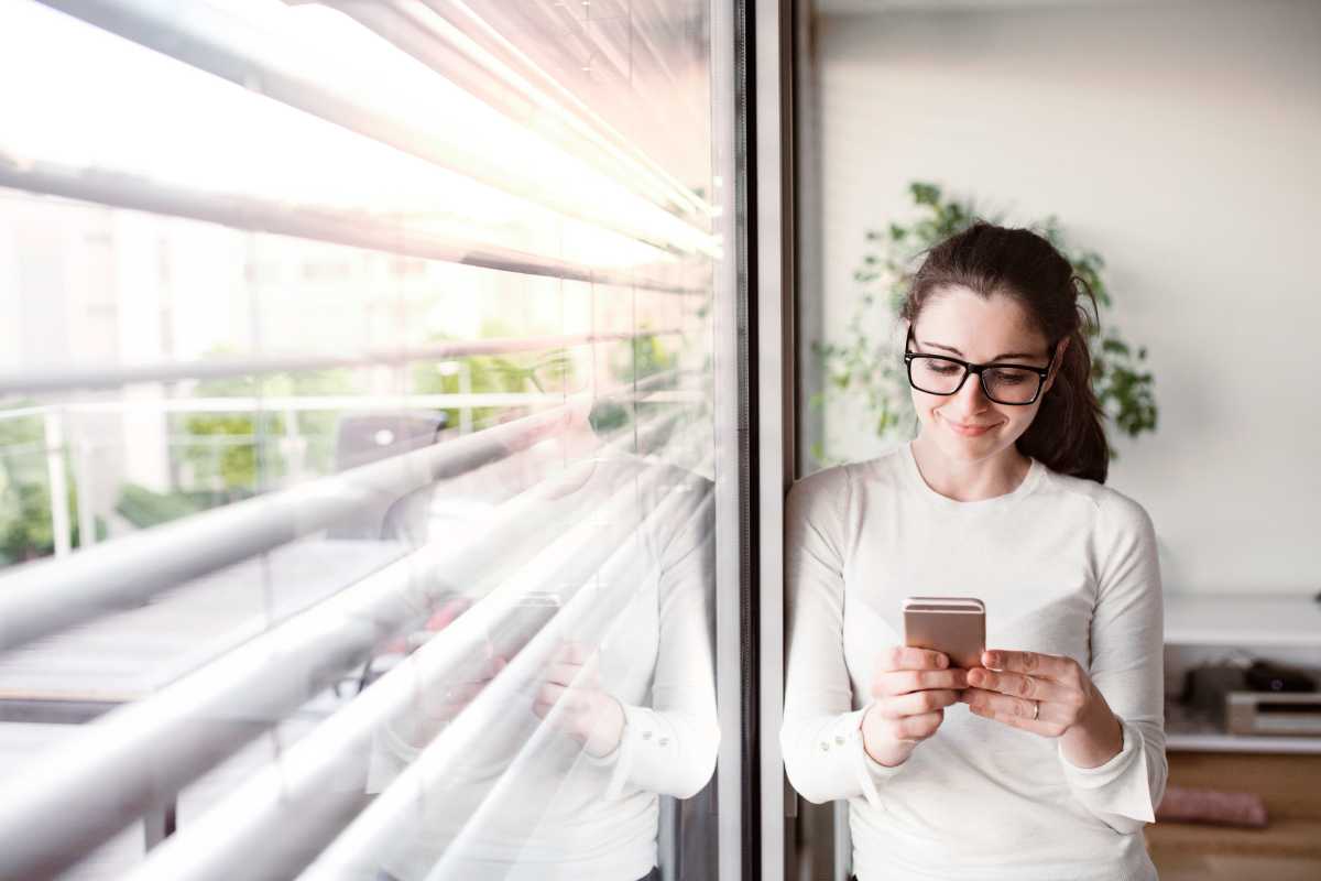 Frau mit Smartphone lehnt am Fenster mit Jalousien